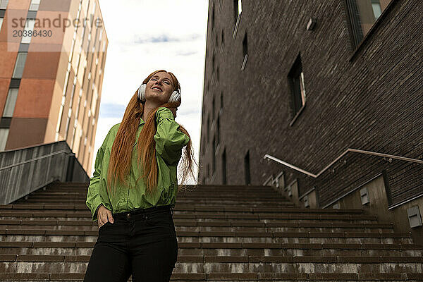 Smiling beautiful young redhead woman listening to music standing on steps