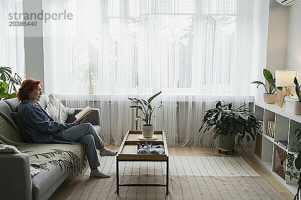 Senior woman reading book sitting on sofa in living room at home