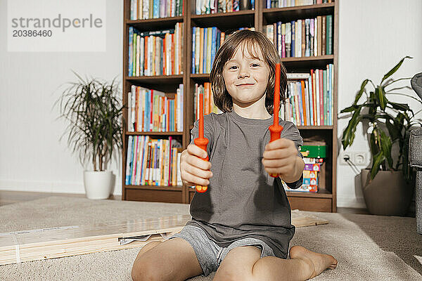 Smiling boy holding screwdrivers sitting in front of bookshelf at home