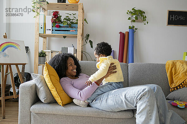 Smiling single mother lying on sofa playing with baby girl in living room at home