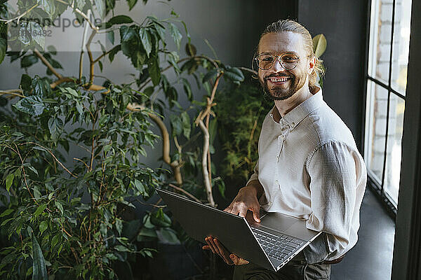 Smiling businessman standing with laptop near plants in office