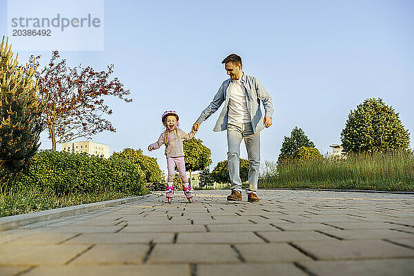 Father holding hand of daughter roller skating on footpath