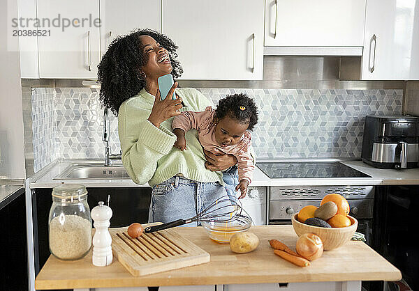 Cheerful single mother carrying daughter talking on mobile phone in kitchen at home