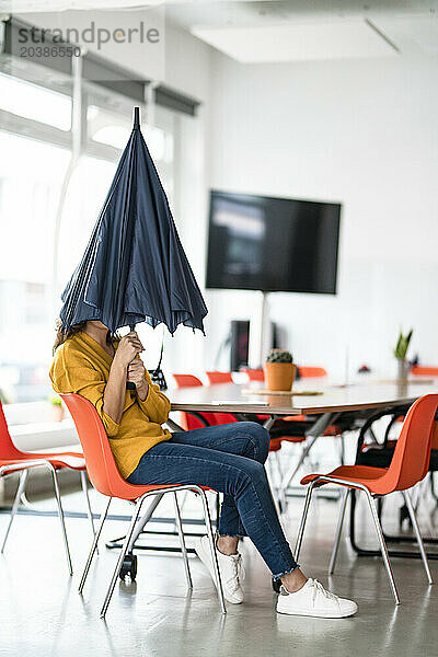 Businesswoman holding folded umbrella covering face in creative office
