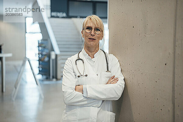 Smiling mature doctor leaning with arms crossed on wall