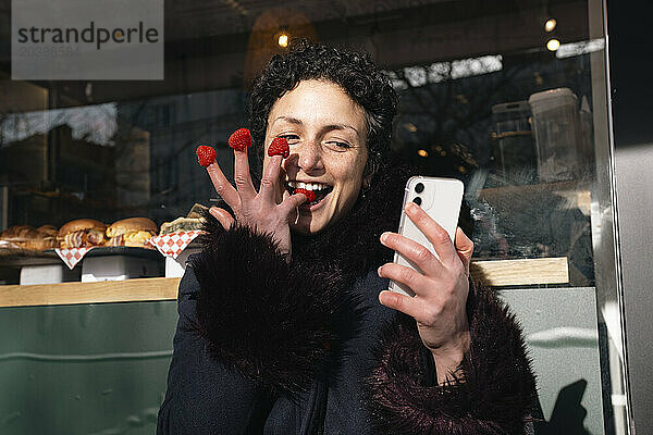 Playful woman eating raspberries on fingers and using smart phone at sidewalk cafe