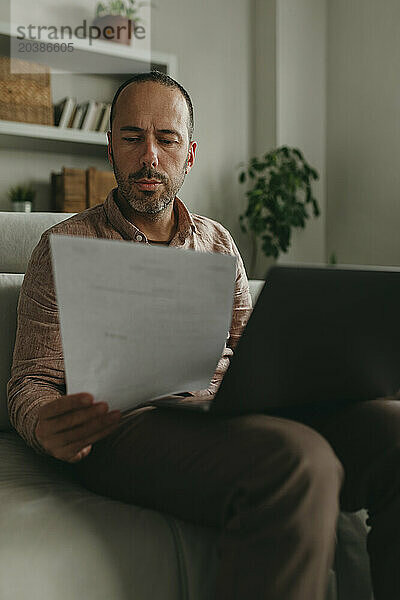 Freelancer examining document near laptop at home office