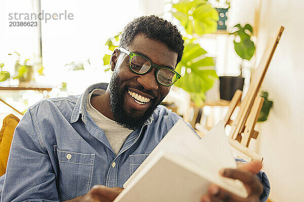 Smiling non-binary person wearing eyeglasses and reading book in living room at home