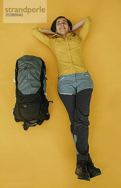 Thoughtful woman lying down near backpack on yellow background