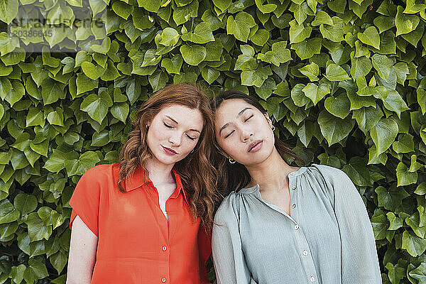 Women with eyes closed leaning on each other near hedge