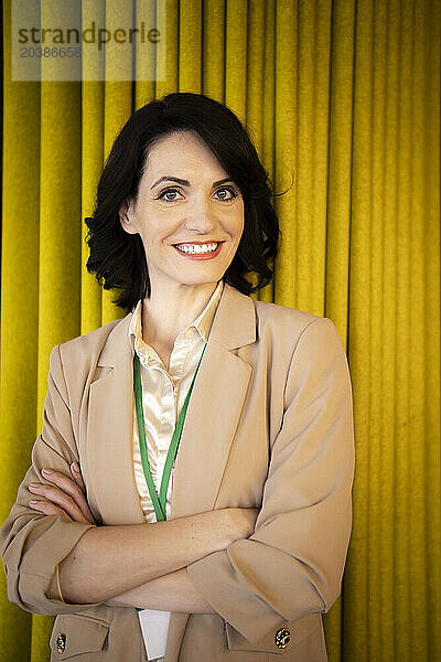 Smiling beautiful businesswoman wearing blazer standing with arms crossed in front of curtain at office