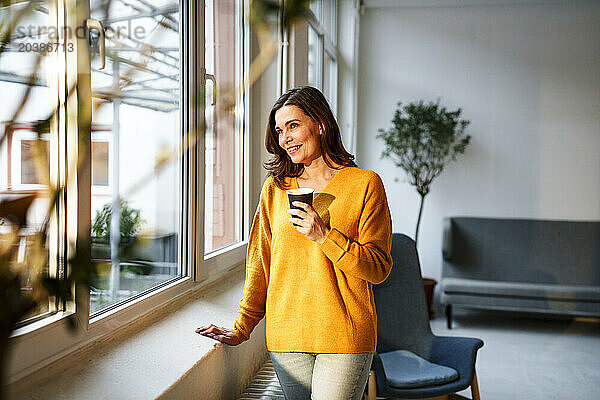 Smiling mature woman holding coffee cup looking out through window at home