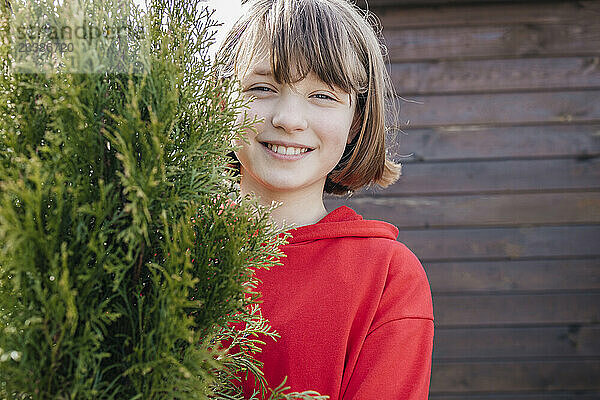 Smiling girl with brown bangs behind Thuja plant