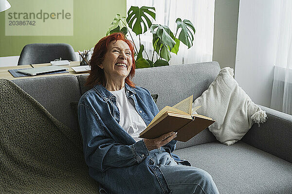 Retired senior woman sitting on sofa with book laughing in living room at home