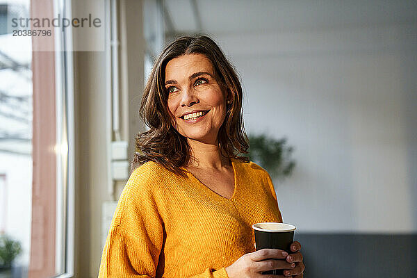 Happy mature woman holding coffee cup looking out through window at home