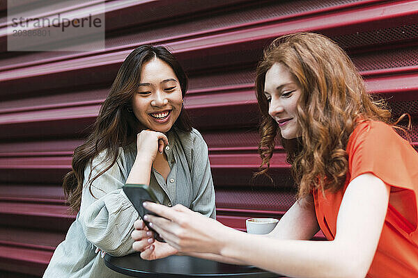 Smiling woman leaning on elbow near friend using smart phone by shutter