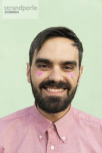 Happy man with pink heart shape stickers on cheeks against mint green background