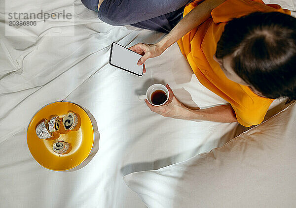 Woman in yellow t-shirt using phone with tea cup and snack lying on bed at home