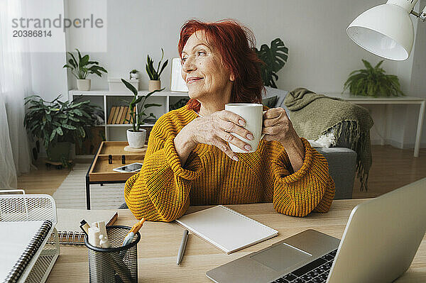 Smiling thoughtful businesswoman with cup sitting at table