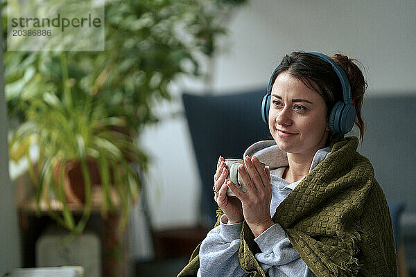 Thoughtful woman with wireless headphones wrapped in blanket holding coffee cup at home