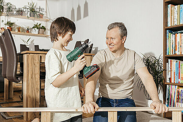 Smiling boy holding power tool by father assembling furniture at home