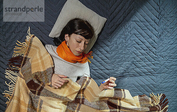 Woman with blanket checking temperature through thermometer on bed at home