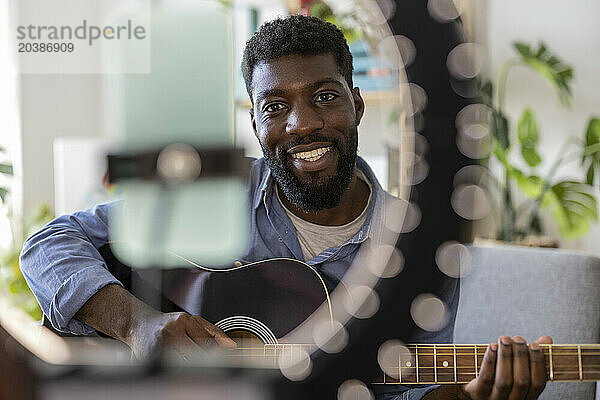 Happy influencer playing guitar and filming through smart phone at home