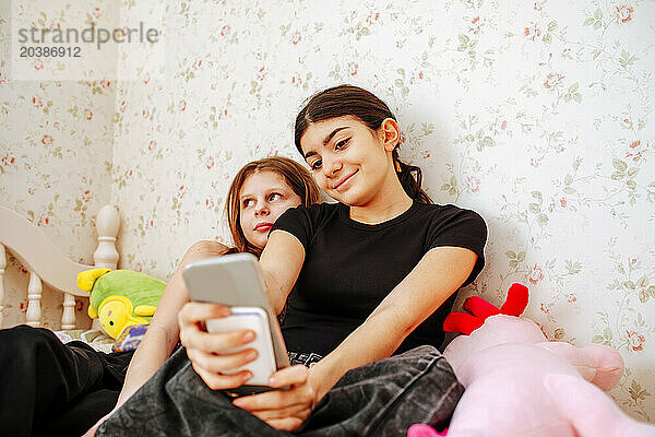 Smiling girl taking selfie through smart phone with friend at home