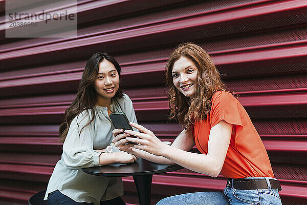 Smiling woman holding smart phone and sitting with friend at sidewalk cafe