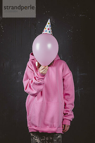 Girl covering face with pink balloon at birthday party in front of black background