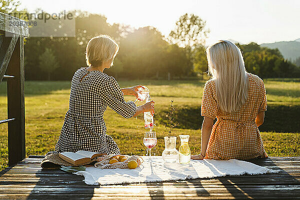 Woman pouring juice by friend and having picnic in park