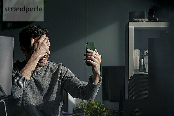 Happy businessman covering face with hand and talking on video call in office