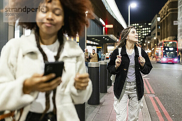 Smiling woman holding backpack and standing near friend using smart phone on street