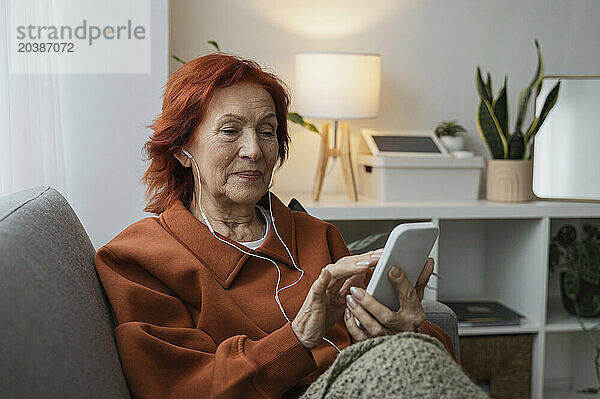 Redhead senior woman using smart phone with headphones sitting on couch in living room