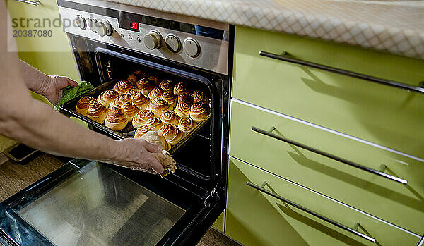 Senior woman taking out tray of sweet dough buns from oven at home