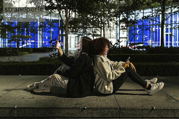 Happy friends sitting back to back and using smart phones at night