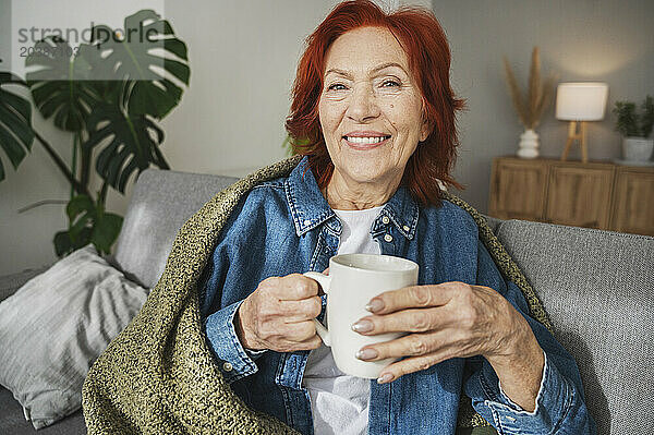 Smiling senior woman sitting under blanket on sofa drinking tea at home