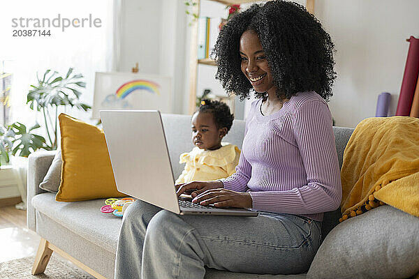 Smiling young mother using laptop working at home sitting by daughter on sofa in living room