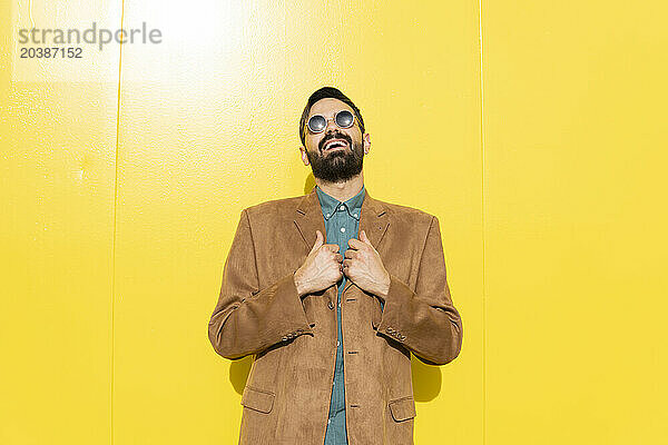 Happy man wearing brown jacket and sunglasses in front of yellow wall