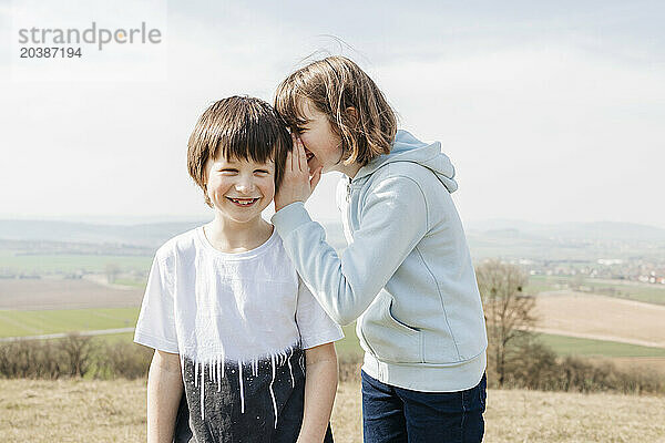 Smiling girl whispering in brother's ear standing on hill