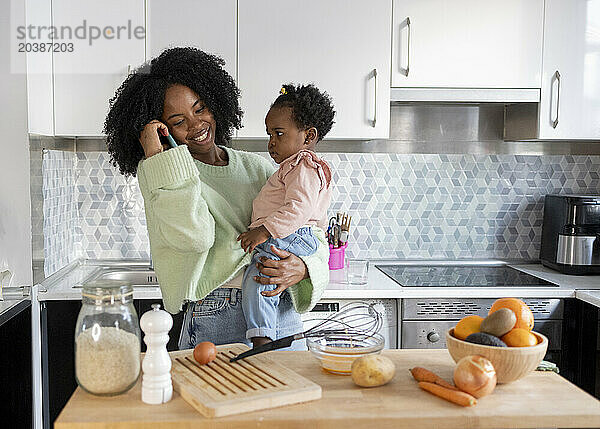 Smiling single mother carrying daughter talking on mobile phone in kitchen at home