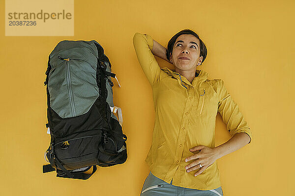 Thoughtful woman lying with backpack on yellow background