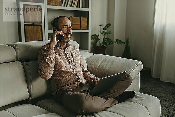 Smiling freelancer sitting with legs crossed and talking on smart phone at home office