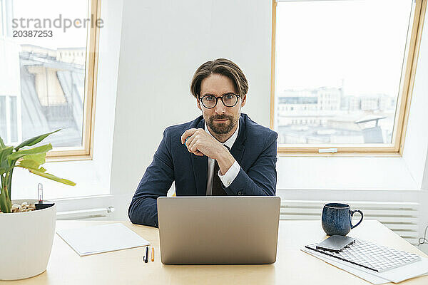 Confident businessman sitting with laptop at desk in window