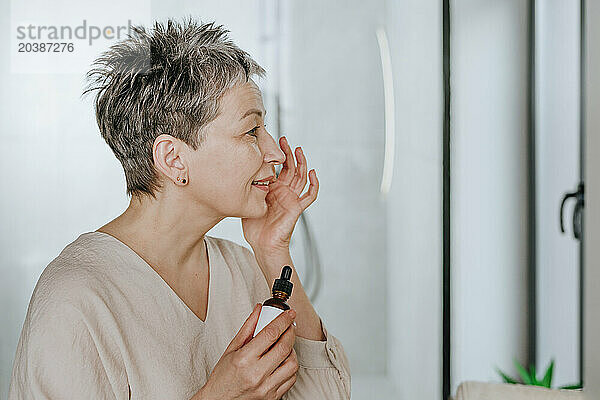 Smiling mature woman applying face serum looking in mirror at home