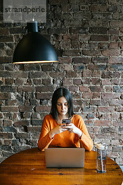 Young woman using smart phone in front of brick wall at cafe