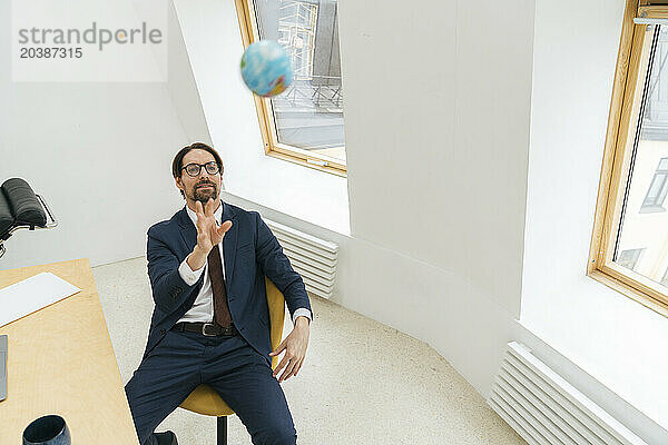 Businessman catching globe sitting by desk and windows in office