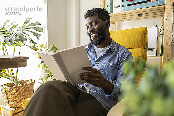 Happy non-binary person reading book on armchair at home