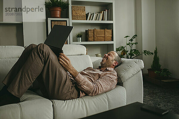 Cheerful man lying down and watching movie on laptop at home