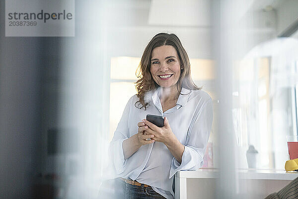 Smiling businesswoman with smart phone sitting at desk in creative office